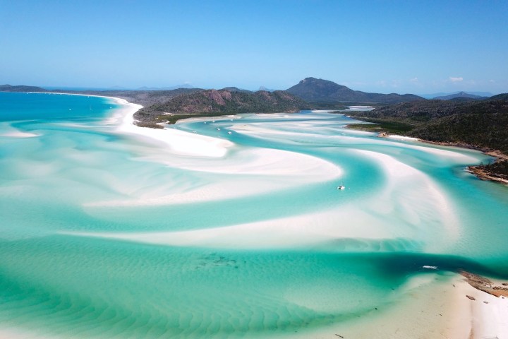 Aerial view of a tropical beach with turquoise water and white sand, surrounded by land and hills.